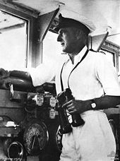 A middle-aged man wearing a white uniform and cap, holding binoculars while looking out a window