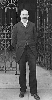 A full height monochrome portrait of a middle-aged white man with a receding hairline and a bushy moustache.