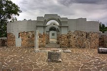 Frontal view through two partially preserved gates of a stone building, with a well in the center of a courtyard in front