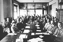 Photo of a caucus meeting showing thirty-five men and a woman, sitting around several long tables with documents laid out before them. As space is cramped and there is not room for everyone at the table, they are seated in two rows.