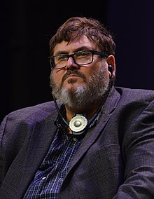 Photo en buste d'un homme brun avec une barbe grisonnante, portant un costume, des lunettes et un appareil auditif de traduction instantanée.