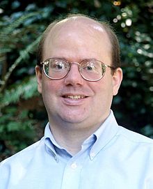 A bespectacled balding man in a blue shirt with the collar unbuttoned looks straight at the viewer.