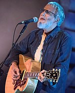 A man in a blue shirt and guitar signing into the microphone