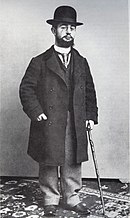 Photo noir et blanc d'un homme à jambe courtes et tronc de taille normale avec une canne. Il porte pantalon à petits carreaux, long manteau, cravate, gilet et chapeau melon. Son visage est marqué par une barbe et un binocle.