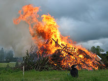 Ein brennender Holzhaufen auf einem Feld. Im Feuer sind Möbelstücke und Europoolpaletten zu erkennen, eine braune Rauchfahne steigt auf. Vor dem Feuer sitzt ein Kind.