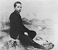 Studio photo of a boy seated in a relaxed posture and wearing a suit, posed in front of a backdrop of scenery.