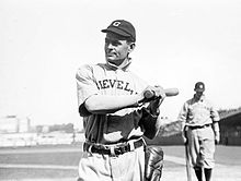 A black-and-white photo of a man in a white baseball uniform with "CLEVELAND" across the chest and a dark baseball cap with "C" on the face in white; he is holding a baseball bat over his left shoulder