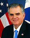 Older man with bushy eyebrows and gray hair in a sui with the US flag behind himt