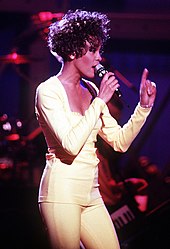 A woman, who wears a light-colored suit, sings and points to the ceiling.