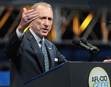 A gray-haired man speaks into two microphones at a podium with a seal reading "AFL–CIO 2009" in the front of it. The man, who is wearing a black suit jacket and blue tie, gestures with his right hand.