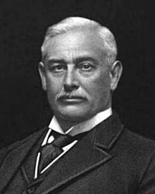 A black-and-white lithographic portrait of a middle-aged, mustached man from the shoulders up, wearing a suit, with a necktie