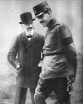 Photographie en noir et blanc de deux hommes debout ; au fond un homme barbu en chapeau ; devant un homme moustachu en uniforme