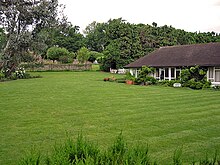 Uma casa branca com um telhado preto ao lado de uma grande jardim bem cuidado. Há várias árvores no fundo da imagem, atrás da casa.