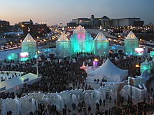 hundreds of spectators in front of the ice castle which is lit blue-green from inside