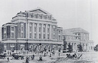 An artistic rendering of the building as built, with two other buildings next to it and people around.