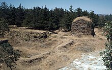 Ruinas cubiertas hierba y maleza con un fondo de bosques de pino bajo. Los escombros de una torre cuadrada está detrás a la derecha, todo lo que queda del Templo de Tohil, con los restos de las paredes del campo de juego de pelota a la izquierda en el primer plano.