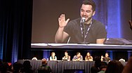 A group of panelists is on a stage with one man being projected onto a large screen above them.