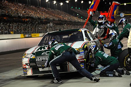 Several men in tracksuits and helmets adjust the tires and refuel the tanks of a race car