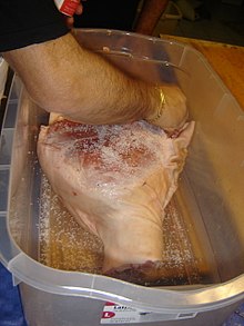 Gros plan sur un bras d'homme en train de frotter de gros sel un énorme jambon placé dans un bassin en plastique transparent.