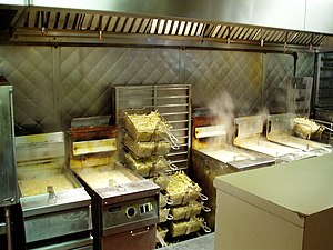 A row of five deep fryers, all steaming and filled with baskets of French fries.