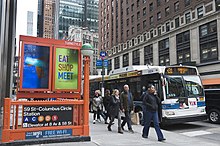 An entrance to the station on 57th Street, which is painted orange. A bus and pedestrians are to the right of the entrance.