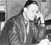 Profile of a white man leaning forward with a large phone handset pressed to his ear. He is wearing a leather jacket over a shirt and tie.