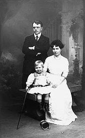 A studio portrait. A young man stands with his arms folded; he has dark hair, and is wearing a dark Edwardian suit, a white shirt with rounded collars, and a dark tie, To his right, a young woman sits on a wooden bench; she has dark, medium length hair, and is wearing a white blouse and a long white skirt. She holds a young fair-haired boy, who is wearing a light tunic with flared skirt and embroidery at the neck, dark boots and short socks. He holds a walking stick in his right hand. An empty paint tin lies on its side near his feet. The background has a colonnade and clouds in the classical romantic style.