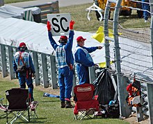 Des commissaires de course agitent simultanément un drapeau jaune et le panneau indiquant l'entrée de la voiture de sécurité en piste.