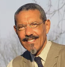 a tan skinned man with narrow glasses smiling in a tight headshot in an sunny outdoor photo