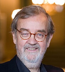 Publicity shot of man with grey hair, white stubble beard and spectacles, smiling.