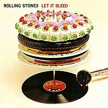 A vinyl record of the album on a record-changer spindle supporting several items stacked on a plate. The topmost item is a cake topped by figurines representing the band.