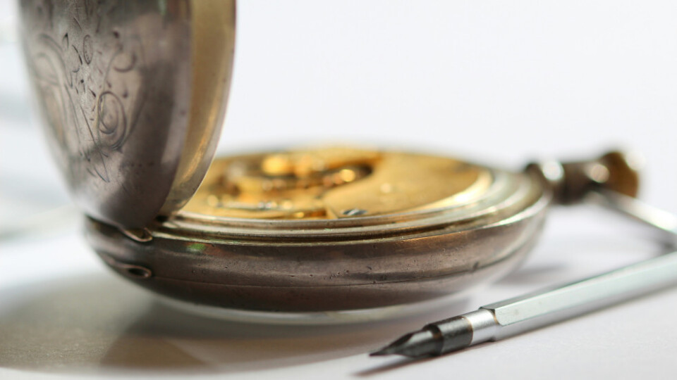 A side view of a pocket watch with a pen lying beside it.