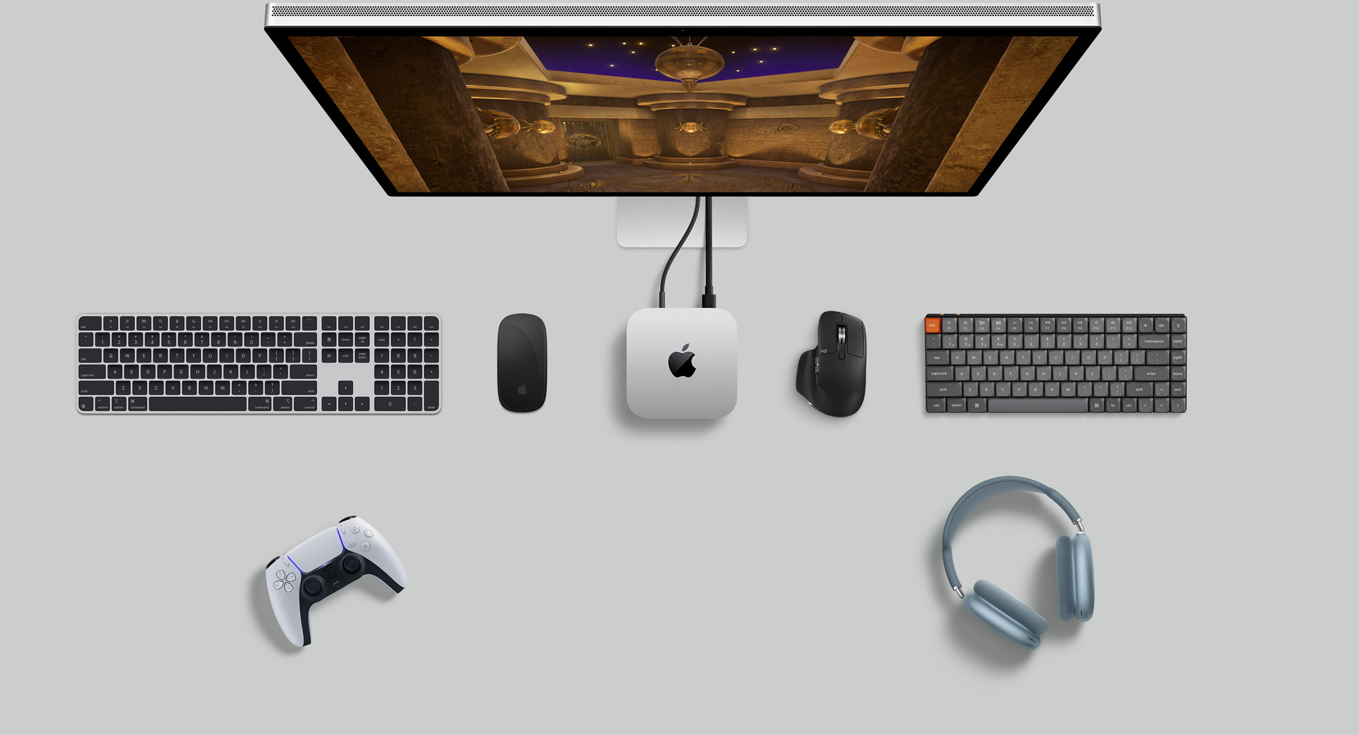 Pohľad zvrchu na strieborný Mac mini zapojený do displeja spoločne na stole s bezdrôtovými klávesnicami, bezdrôtovou myšou, herným ovládačom a slúchadlami AirPods Max