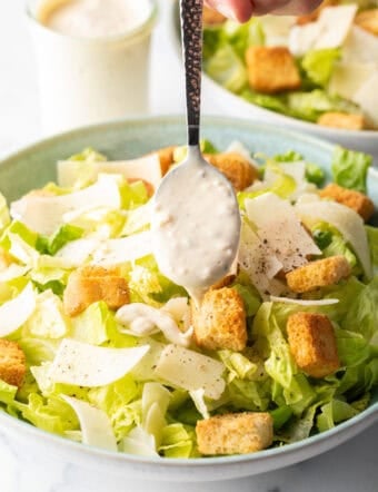 Spooning homemade Caesar dressing over a salad made with chopped romaine, croutons, and parmesan cheese.