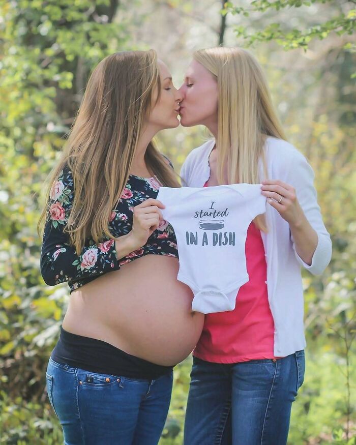 Two women outdoors, one pregnant, share a kiss and hold a baby outfit with text.