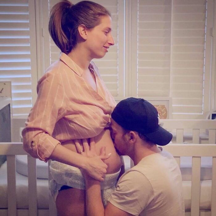 Pregnant woman standing, partner kneeling and kissing her belly in a heartfelt moment.