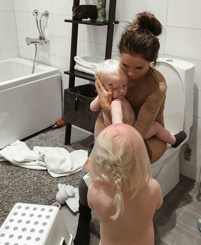 Woman in a bathroom holding a child, another child stands nearby, capturing a real-life parenting moment.