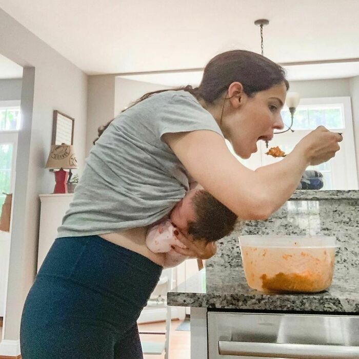 Woman multitasking in a kitchen, holding a baby and eating from a container; real-life moment.