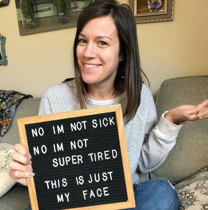 Woman smiling, holding a sign that reads, "No I'm not sick, no I'm not super tired, this is just my face."