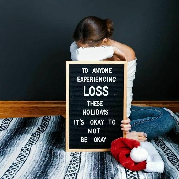 Woman sitting on the floor, holding a sign about experiencing loss during holidays, emphasizing it's okay to not be okay.