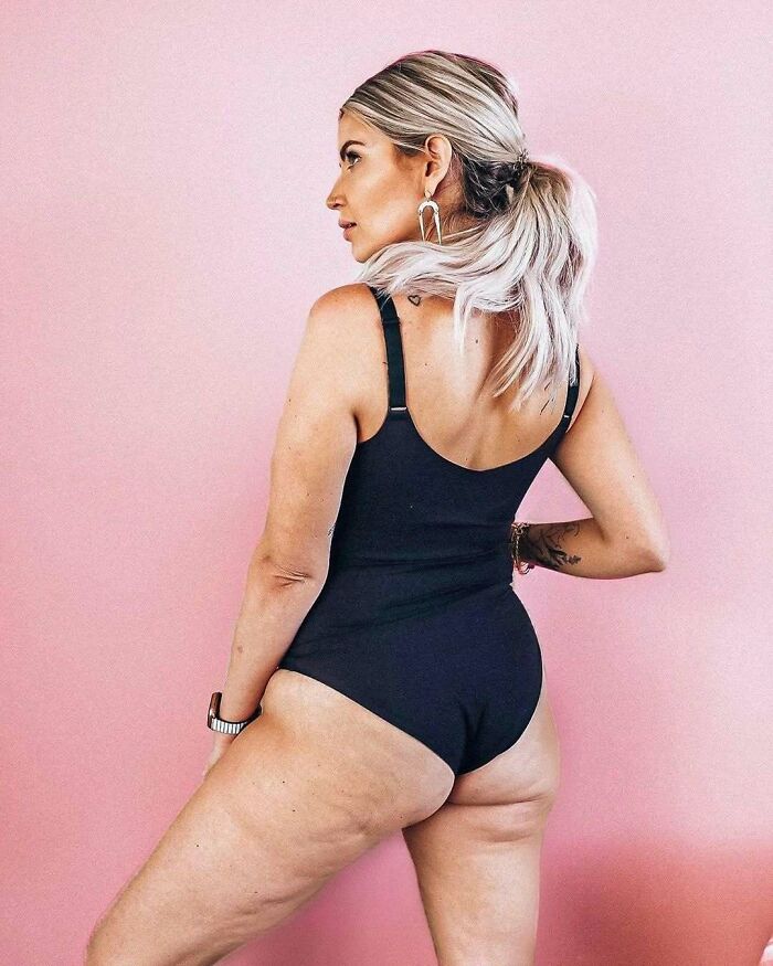 Woman in black swimsuit posing confidently against a pink background, showcasing body positivity.