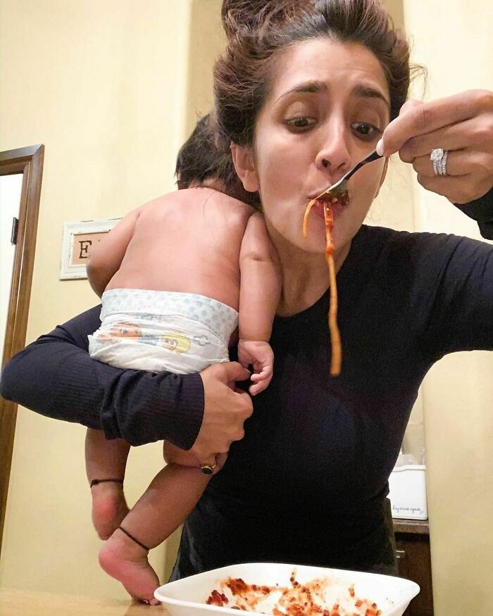 Woman balancing a baby on her arm while eating spaghetti, capturing a real-life multitasking moment.