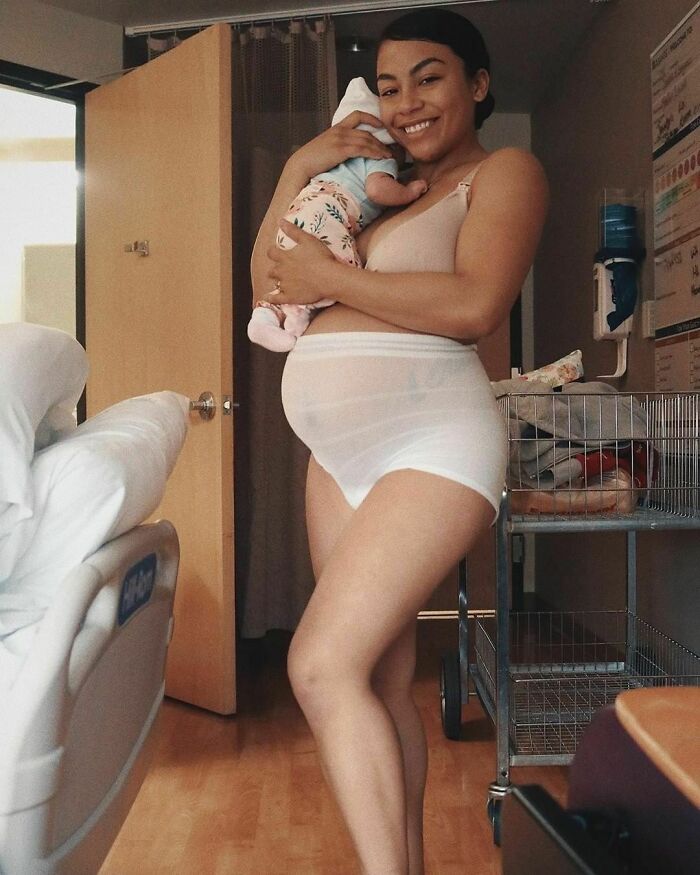 Woman holding newborn in a hospital room, smiling and wearing postpartum underwear, showcasing real-life moments.