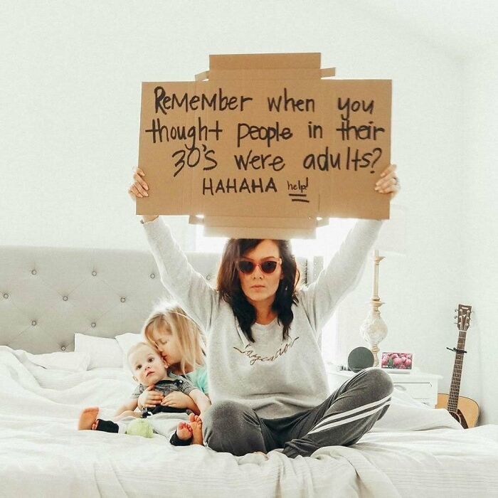 Woman on a bed with kids, holding a funny sign about adulthood, representing women in real-life moments.