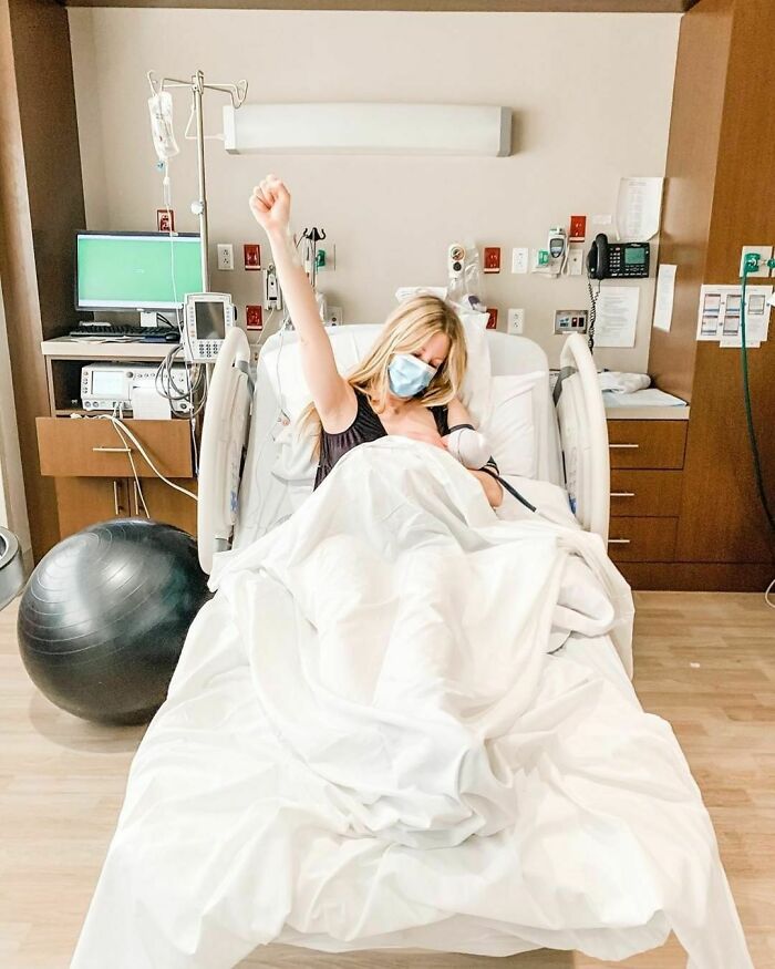 Woman in a hospital bed after childbirth, wearing a mask, raising her fist in triumph, embodying Women-In-Real-Life-Pics.