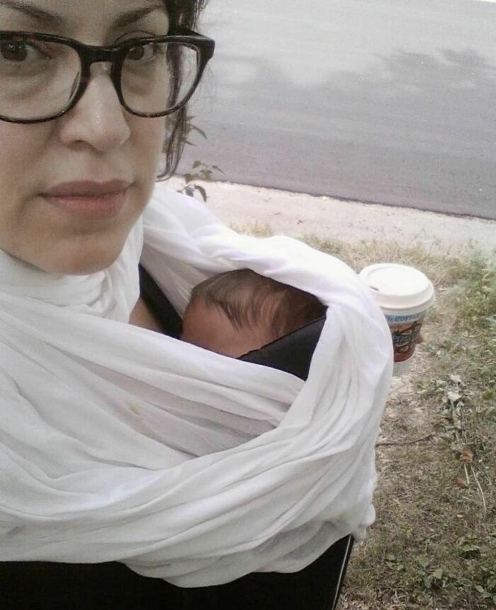 Woman carrying a baby in a wrap, holding a coffee cup, representing women in real life moments outdoors.