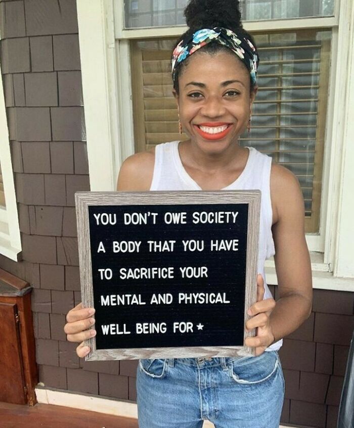 Woman smiling, holding a sign promoting self-acceptance and well-being on a porch.