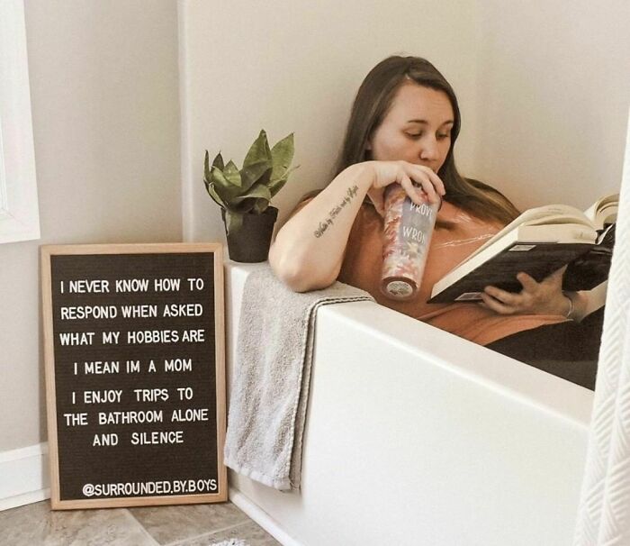 Woman relaxing in a bathtub, reading a book, and drinking from a cup next to a letter board with a humorous message.