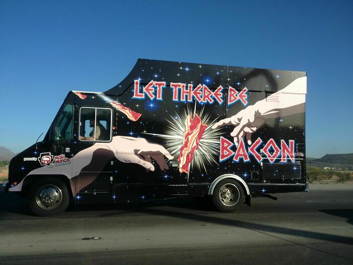 Food truck with cosmic bacon art and slogan, "Let There Be Bacon," driving on the road.