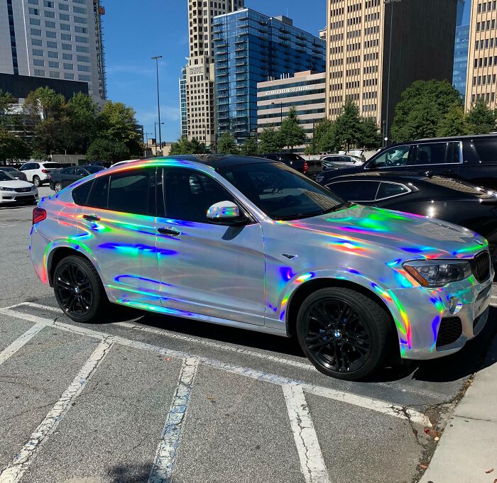 Iridescent car in city parking, reflecting rainbow colors under sunlight.